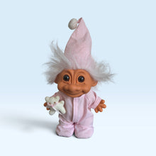 A russ troll figurine wearing pink pinstriped pajamas and a matching pointed hat with a pom-pom, holding a small white stuffed animal. The figurine has brown skin, large dark eyes, and messy white hair, set against a light blue gradient background.